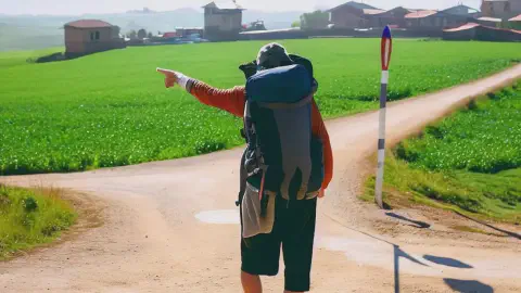 Pilgrim At Crossroads On Camino De Santiago Pilgrim At Crossroads On Camino De Santiago Walking Away Pointing Left Sunny Day Green Fields Pathway Villages
