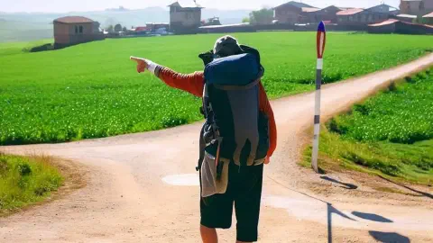 Pilgrim At Crossroads On Camino De Santiago Pilgrim At Crossroads On Camino De Santiago Walking Away Pointing Left Sunny Day Green Fields Pathway Villages